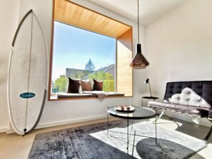 Quarto moderno com uma janela grande, um sofá estofado preto, uma mesa de centro redonda e uma prancha de surf encostada na parede.