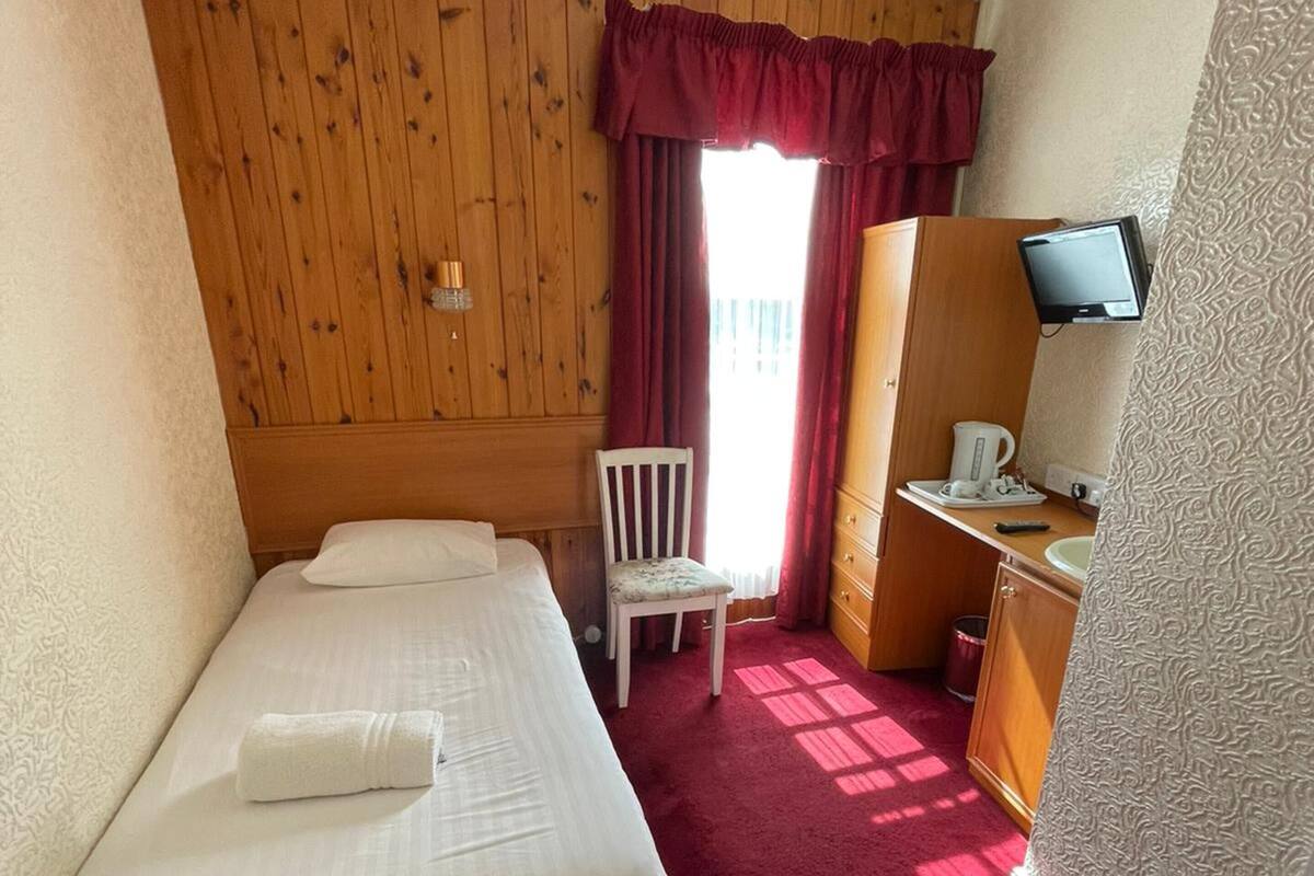 Ein Hotelzimmer mit einem Einzelbett, einem Sessel, einem Schreibtisch mit Fernseher und einem Kleiderschrank.