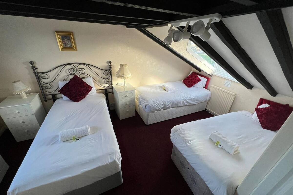 Ein Schlafzimmer mit zwei Betten, jedes mit einem roten Kissen und weißer Bettwäsche. Ein Nachttisch mit Lampe und ein kleiner Tisch mit Lampe sind vorhanden. Über den Betten hängt ein gerahmtes Bild.