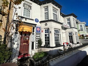 Un immeuble d’angle à la façade blanche, avec une porte rouge et une enseigne bleue indiquant qu’il s’agit d’un hôtel privé.