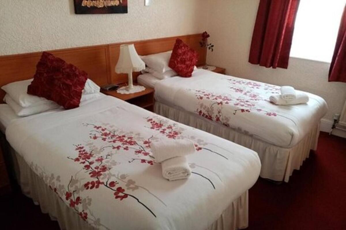 Ein Hotelzimmer mit zwei Betten, jeweils mit floralem Überwurf und roten Kissen. Ein Nachttisch mit Lampe und ein kleiner Tisch mit einer roten Blumen Dekoration.