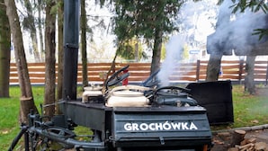 Un equipo de cocina portátil con la marca GROCHOWKA, que presenta una chimenea que emite humo, ubicado al aire libre con árboles y una valla de fondo.