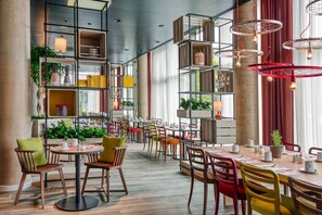 A modern restaurant with wooden tables and chairs, shelves with books and plants, and red hanging lights.
