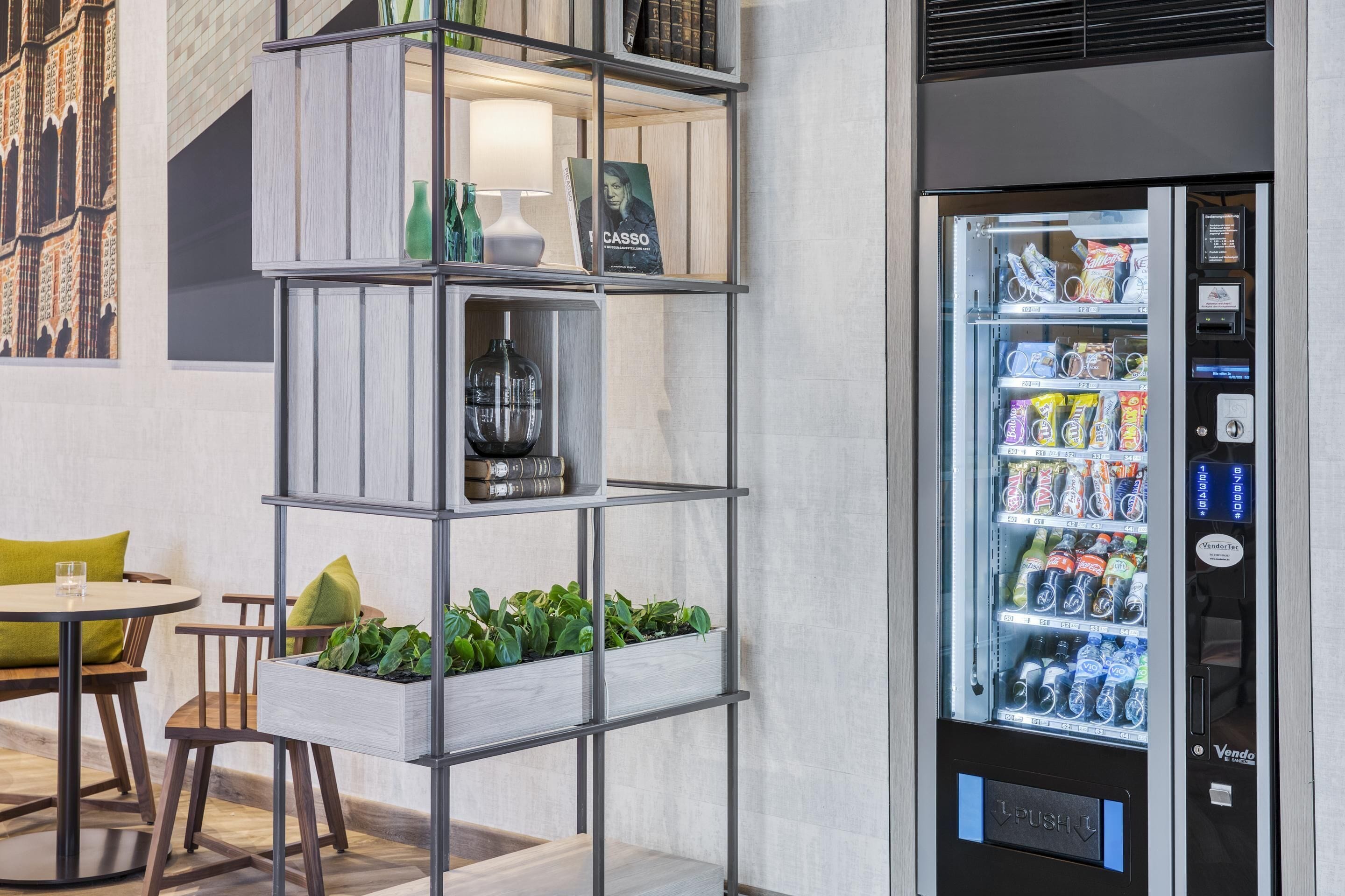 Un interior moderno con una máquina expendedora llena de bebidas y aperitivos, una estantería de madera con objetos decorativos y una pequeña mesa con sillas.