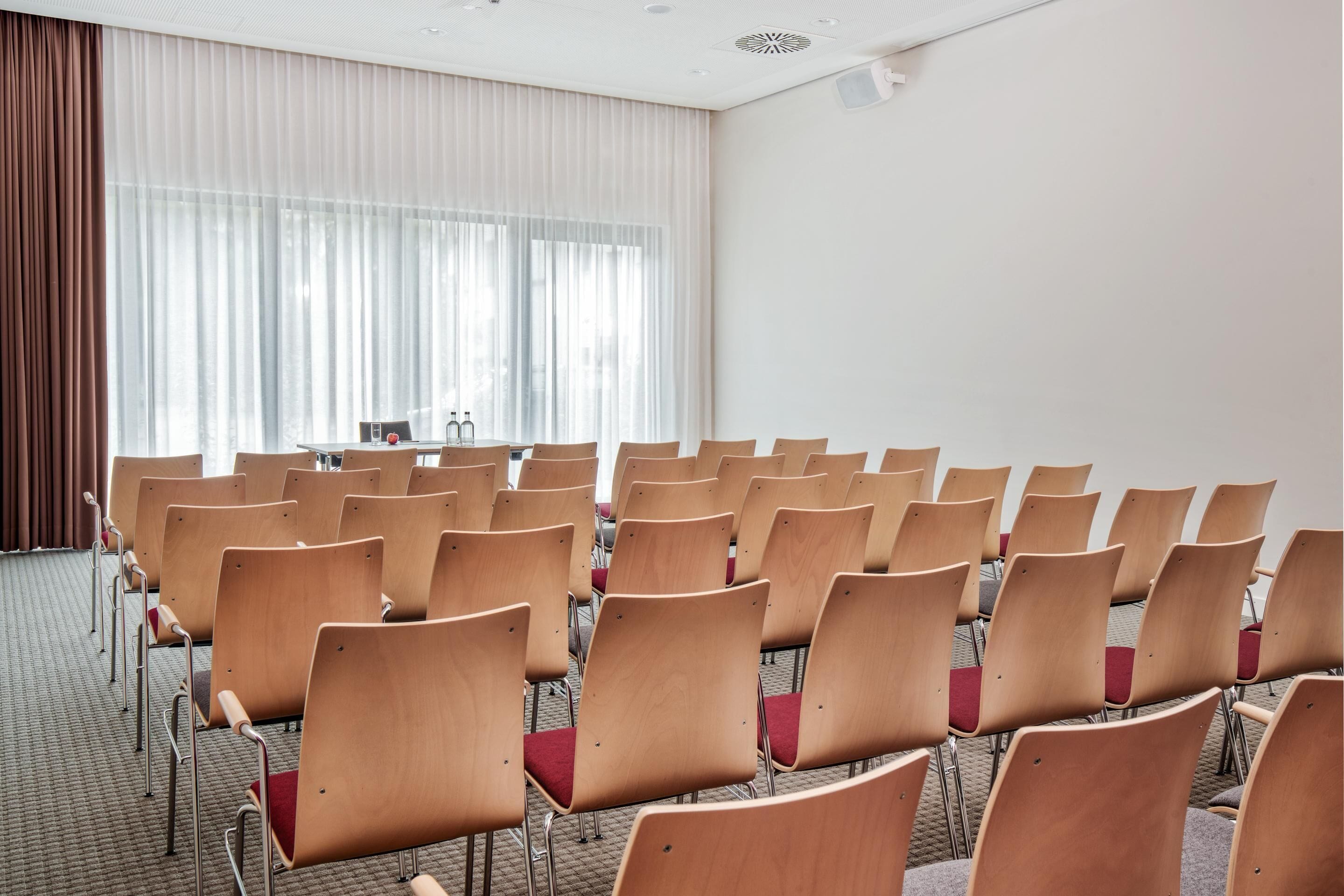 A conference room with rows of wooden chairs and a large window with sheer curtains.