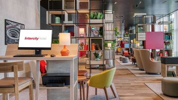Een moderne hotellobby met een receptiebalie, een zithoek en diverse planten en boekenkasten.