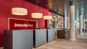 A reception area with a red wall and a sign that reads "Intercity Hotel". There are three reception desks, a wooden floor, and a ceiling with recessed lighting. - IntercityHotel Saarbrücken (Saarbrücken)