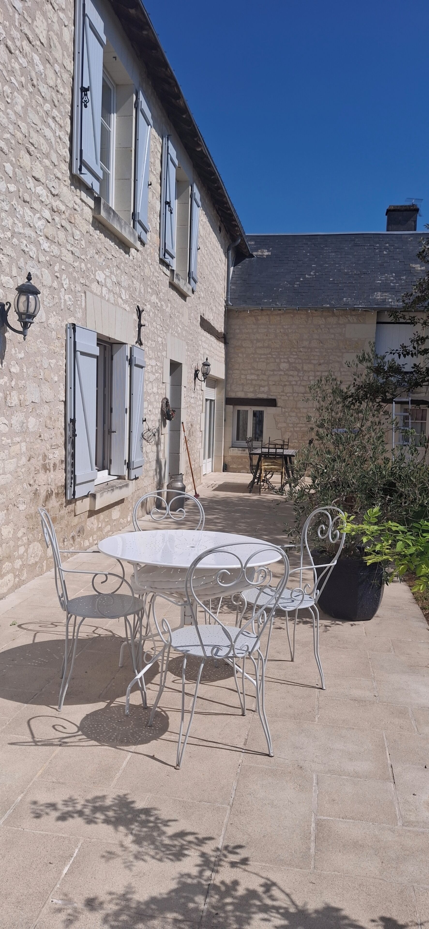 Une cour avec une table ronde et des chaises, un bâtiment en pierre avec des volets, et un ciel bleu dégagé.