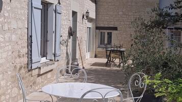 Une cour avec une table ronde et des chaises, un bâtiment en pierre avec des volets, et un ciel bleu dégagé.