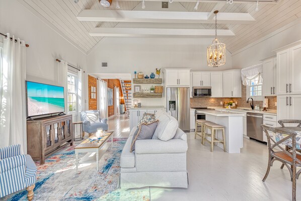 A spacious living area with a large TV, a sofa, and a dining table with chairs. There is a kitchen with white cabinets and a hanging light fixture. - WANDERLUST ~ Stunning 3 Bed, 3 Bath Retreat in Peaceful Neighborhood w/ Pool! (Key West)