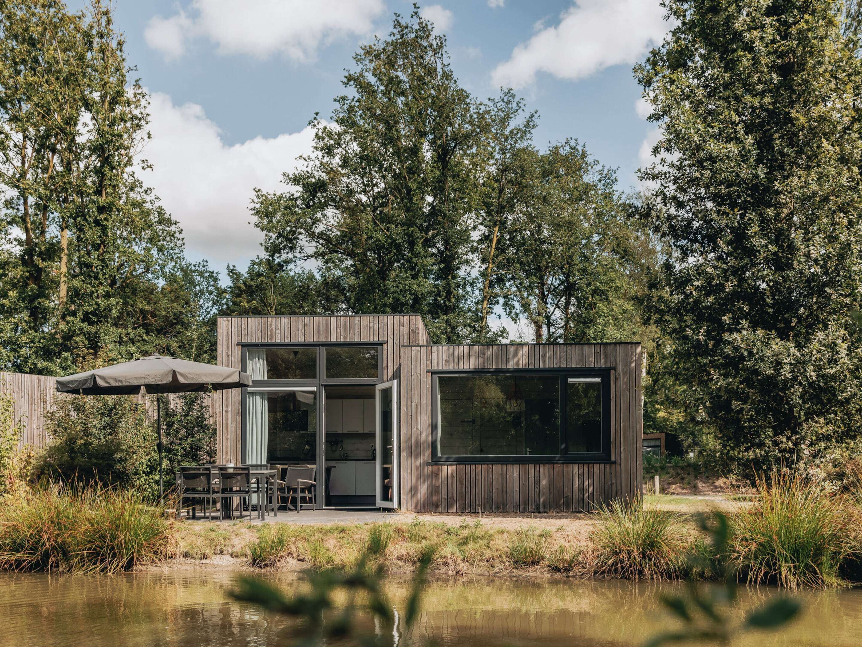 A modern wooden cabin with a glass door entrance, a patio with outdoor furniture, and a view of a lake surrounded by trees.