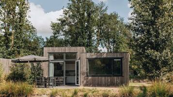 Een moderne houten cabin met een glazen deur, een terras met tuinmeubilair en uitzicht op een meer omringd door bomen.