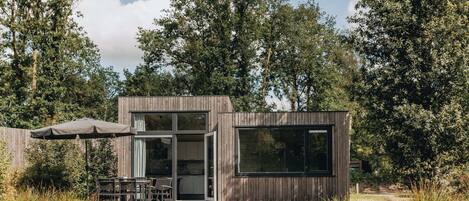 A modern wooden cabin with a glass door entrance, a patio with outdoor furniture, and a view of a lake surrounded by trees.