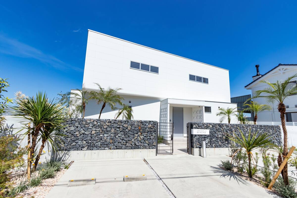 A modern two-story building with a stone wall entrance, palm trees, and a clear blue sky.