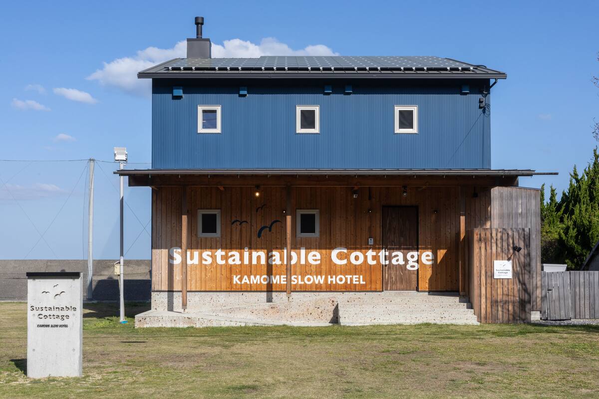 A two-story building with a blue exterior and wooden lower section, labeled as "Sustainable Cottage" and "Kamome Slow Hotel".