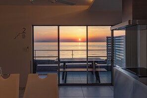Moderni huone, jossa on suuri liukuovi, joka johtaa parvekkeelle, jolta avautuu upea näkymä auringonlaskuun meren yllä.