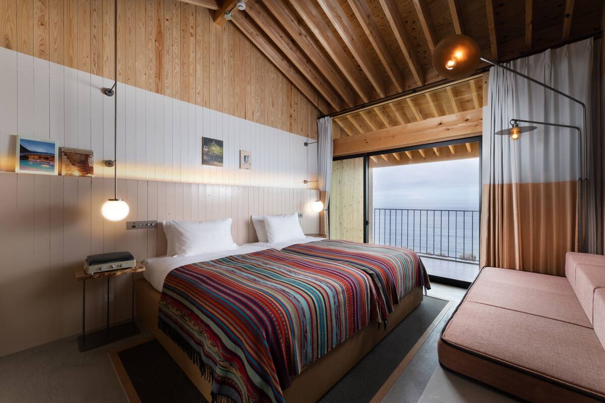 A bedroom with a large bed, wooden ceiling beams, a view to the outside, and a small table with a lamp.