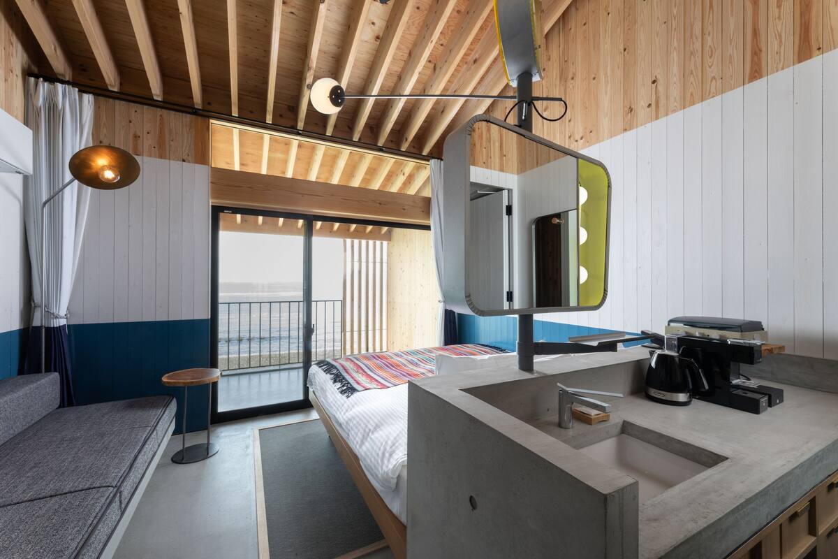 A modern hotel room with a large bed, a sitting area, a bathroom with a sink and mirror, and a balcony with a view.