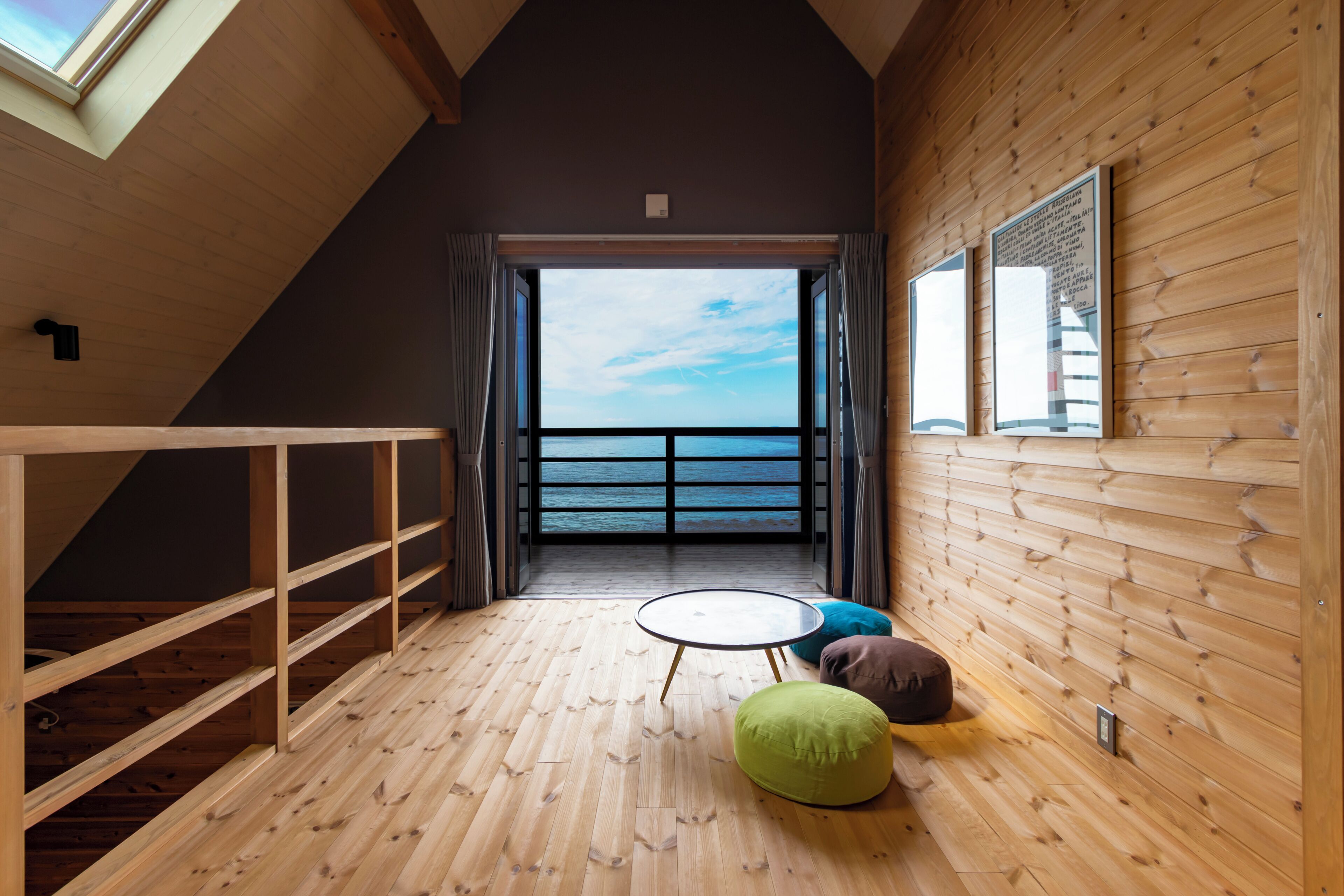 A room with a wooden floor, a balcony with a view of the sea, and a small round table with a cushion.