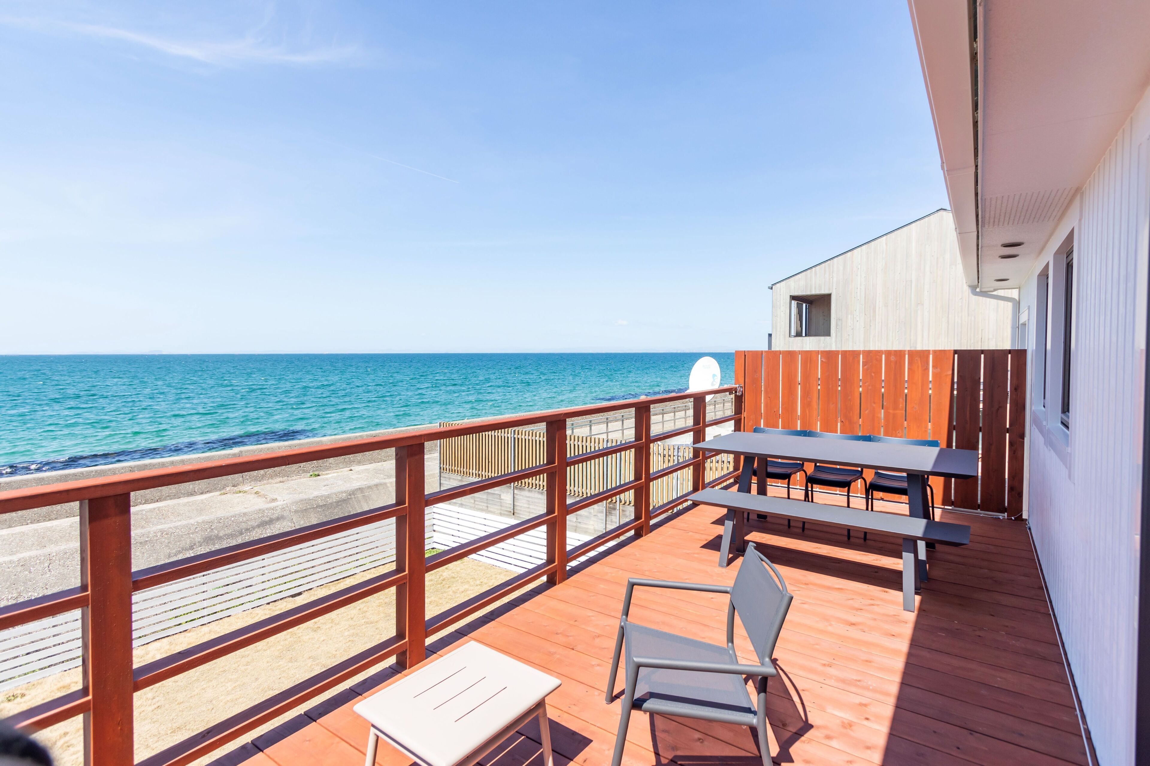 A wooden deck with a table and chairs, overlooking a beach and the sea.