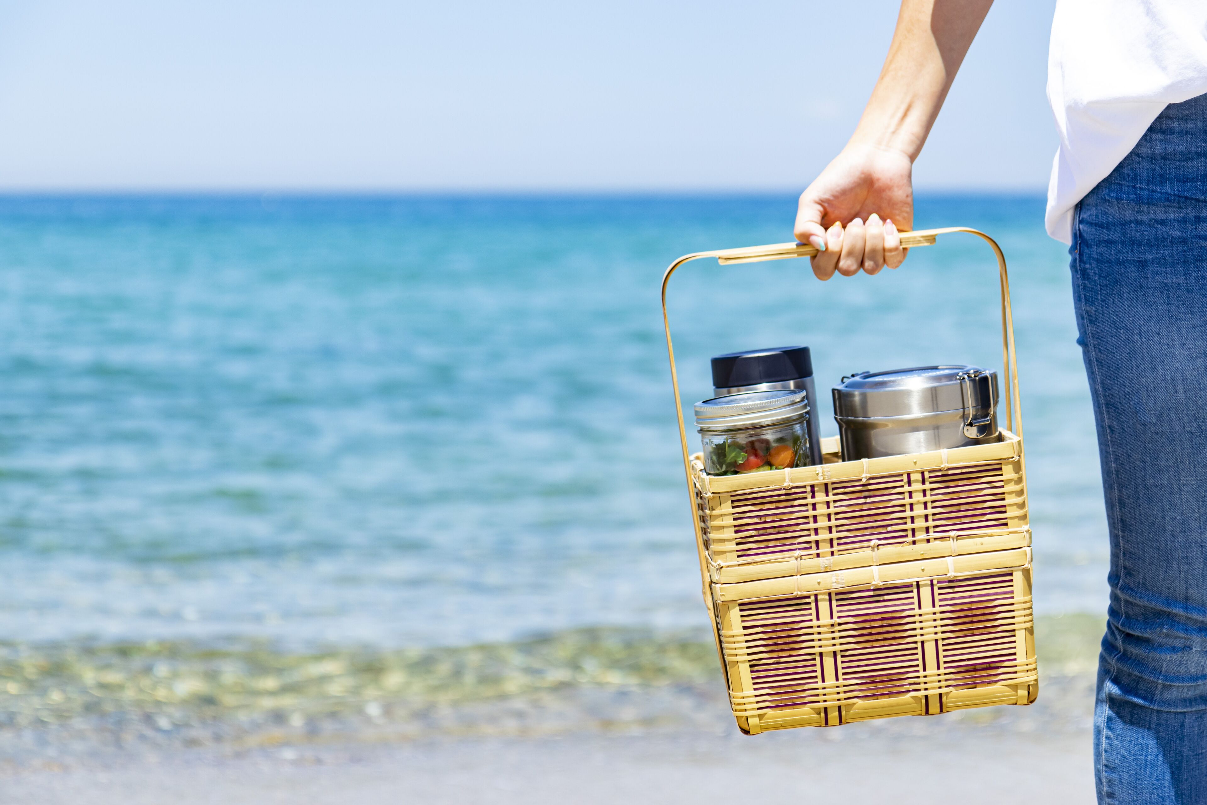 Una persona sostiene una cesta de mimbre con comida y un termo cerca de la playa.