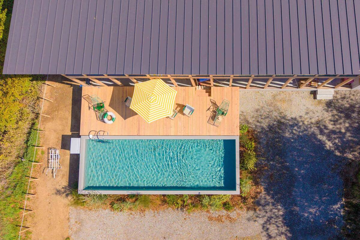 An aerial view of a swimming pool with a deck, surrounded by greenery and a covered area with seating.