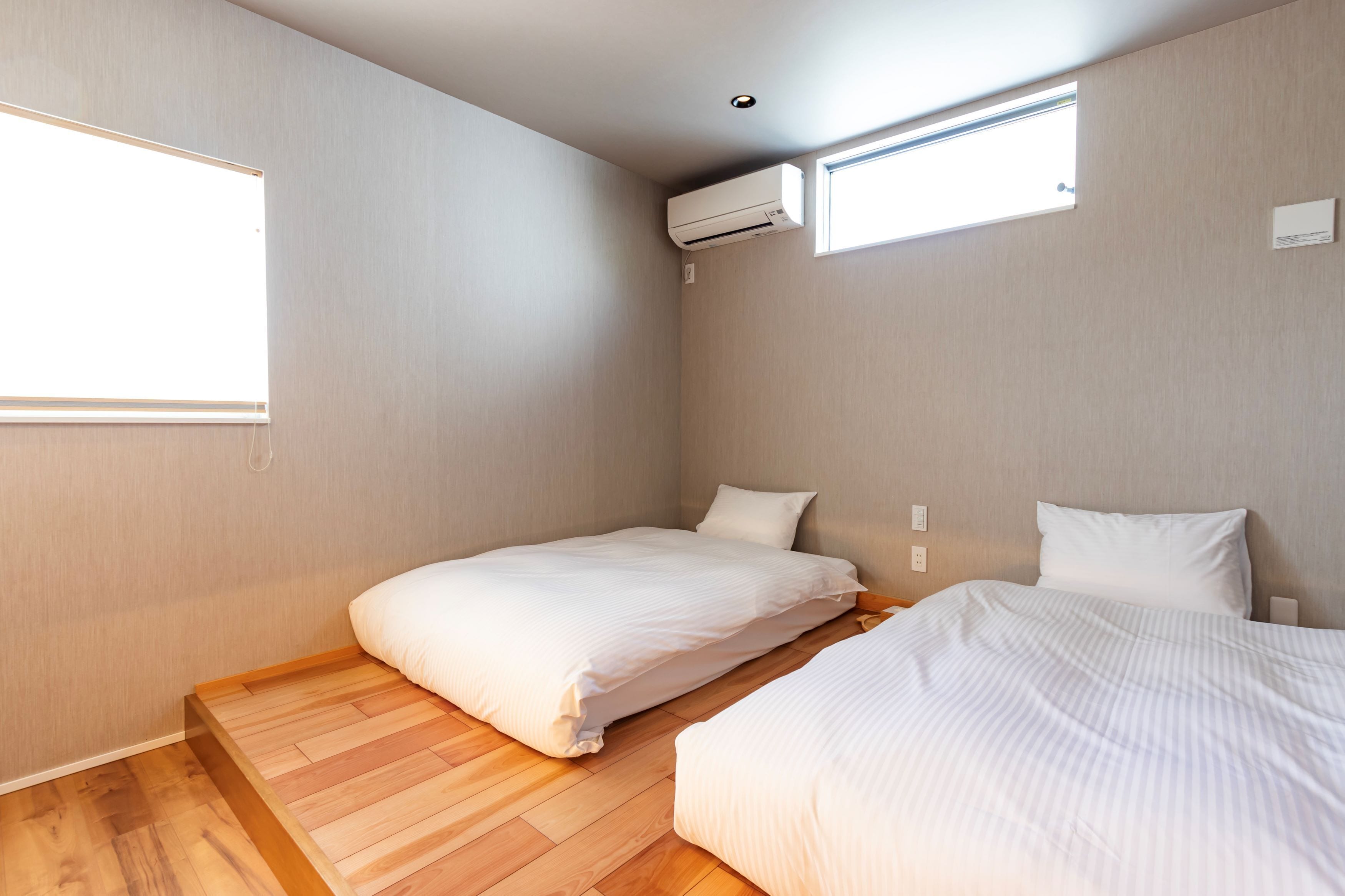 A small, modern room with two beds, a wooden floor, and a window providing natural light.