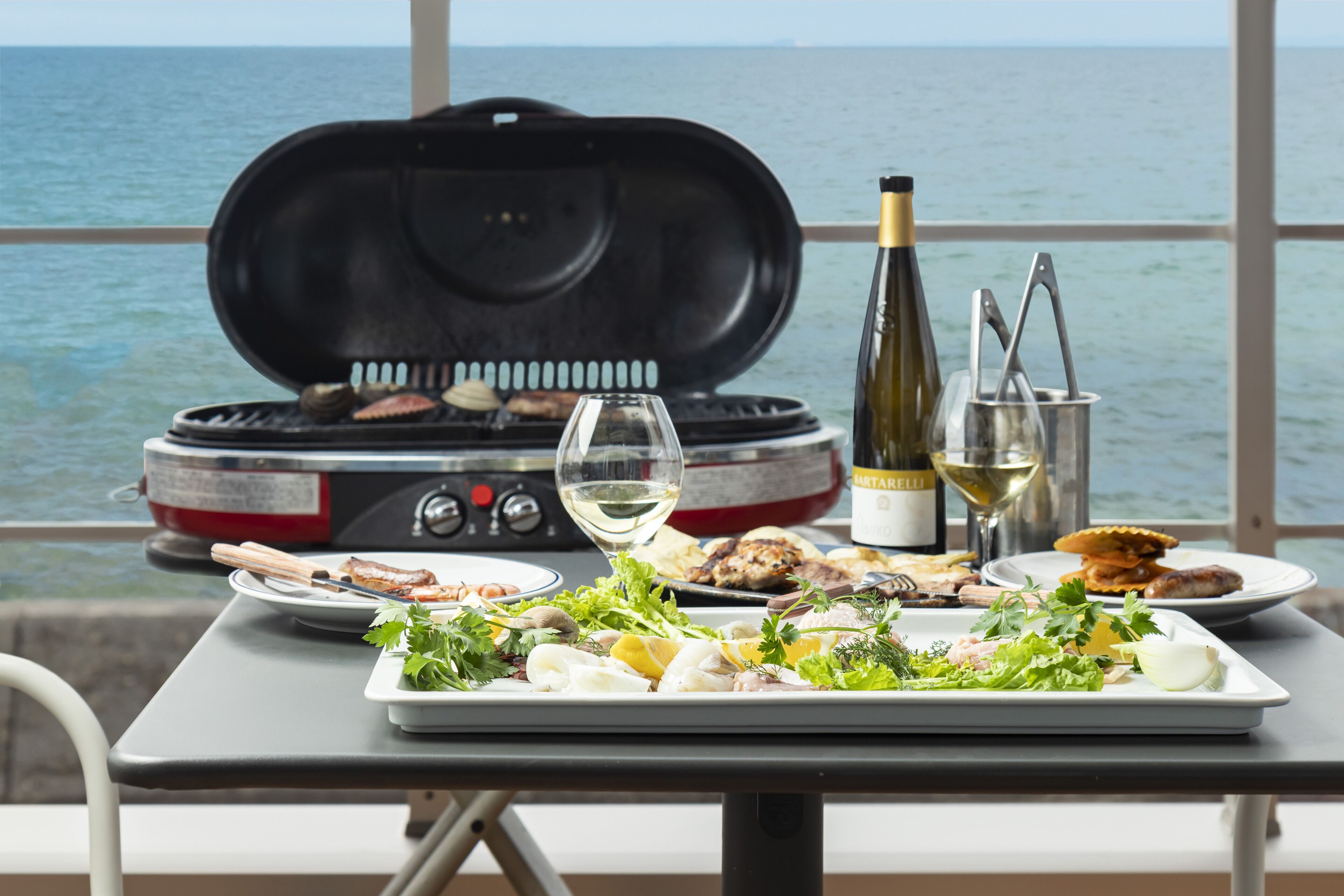A table with a variety of seafood and grilled meats, a wine bottle, and a grill with food being cooked.