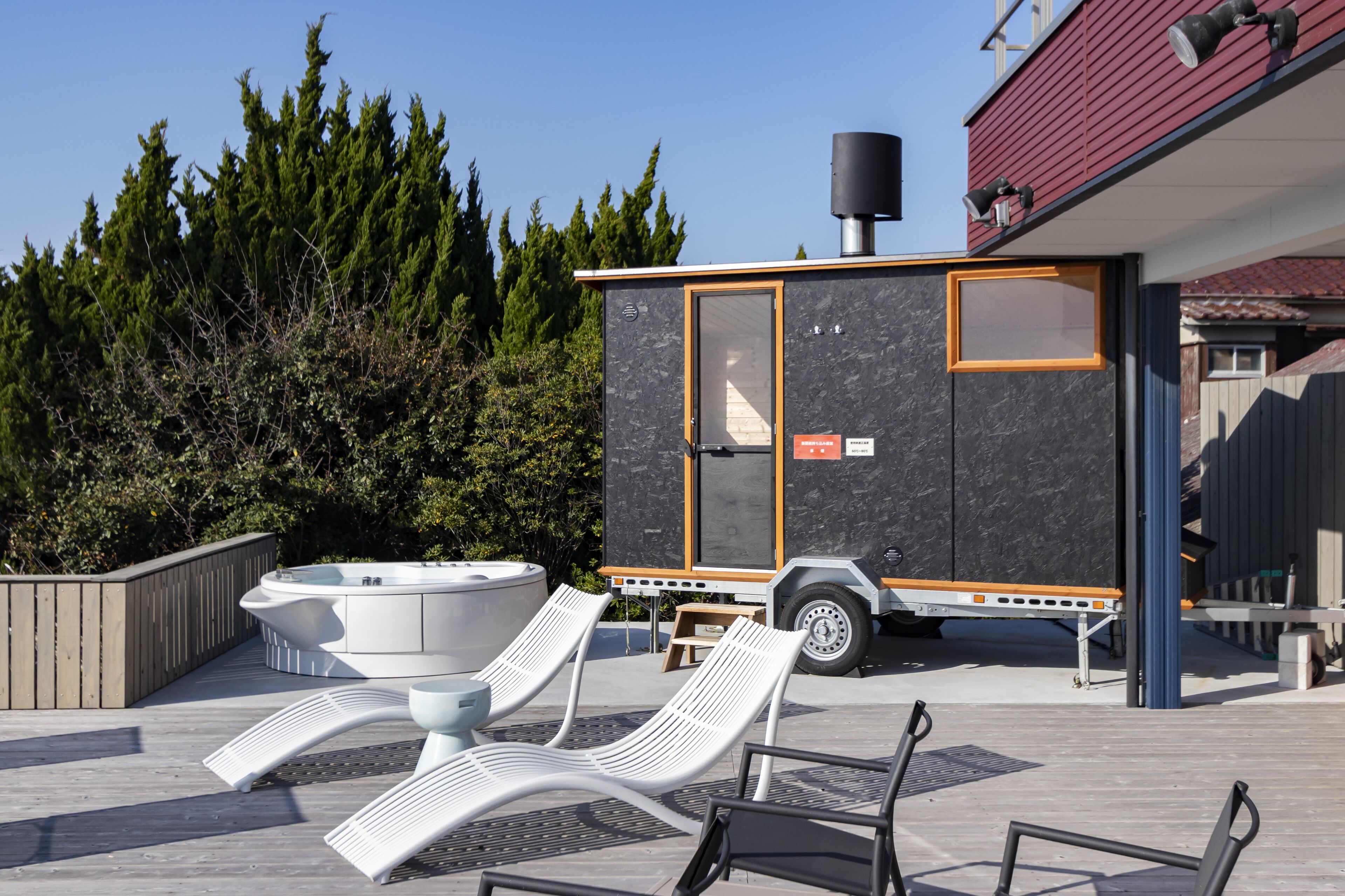 A hot tub and lounge chairs on a wooden deck, a portable sauna, and a building with a red roof and a black chimney.