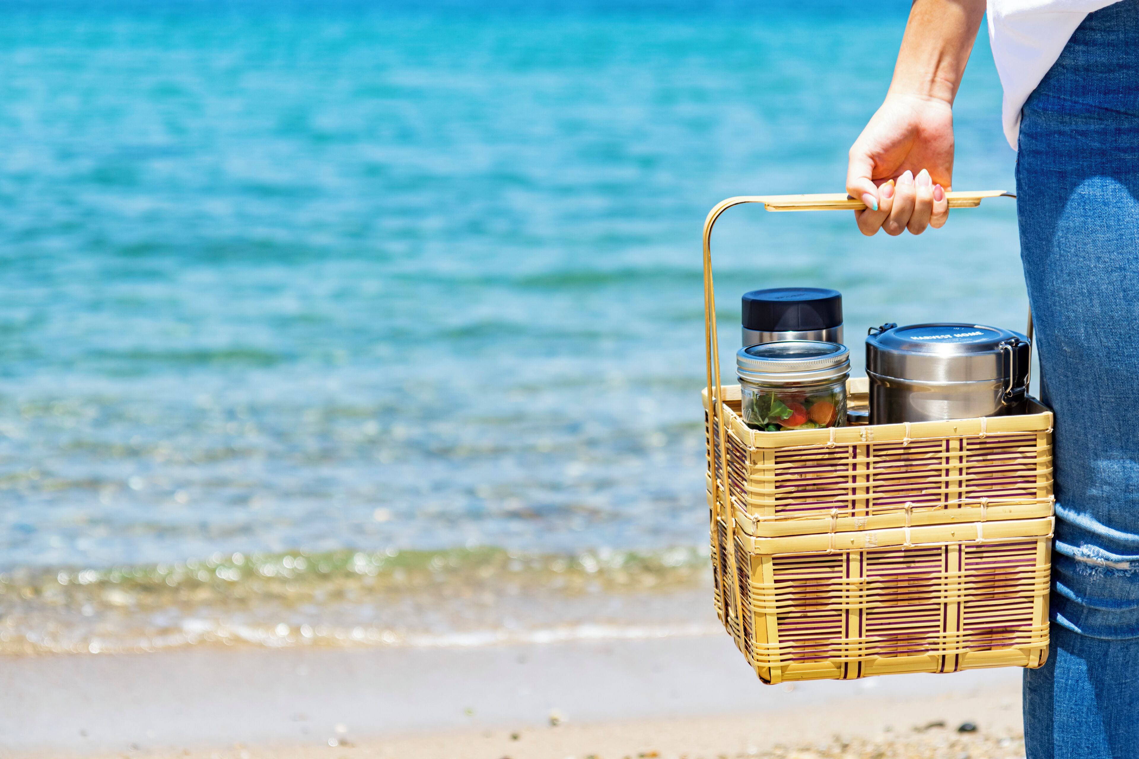 A person holding a wicker basket with a metal container and a thermos, standing by a beach with clear water.