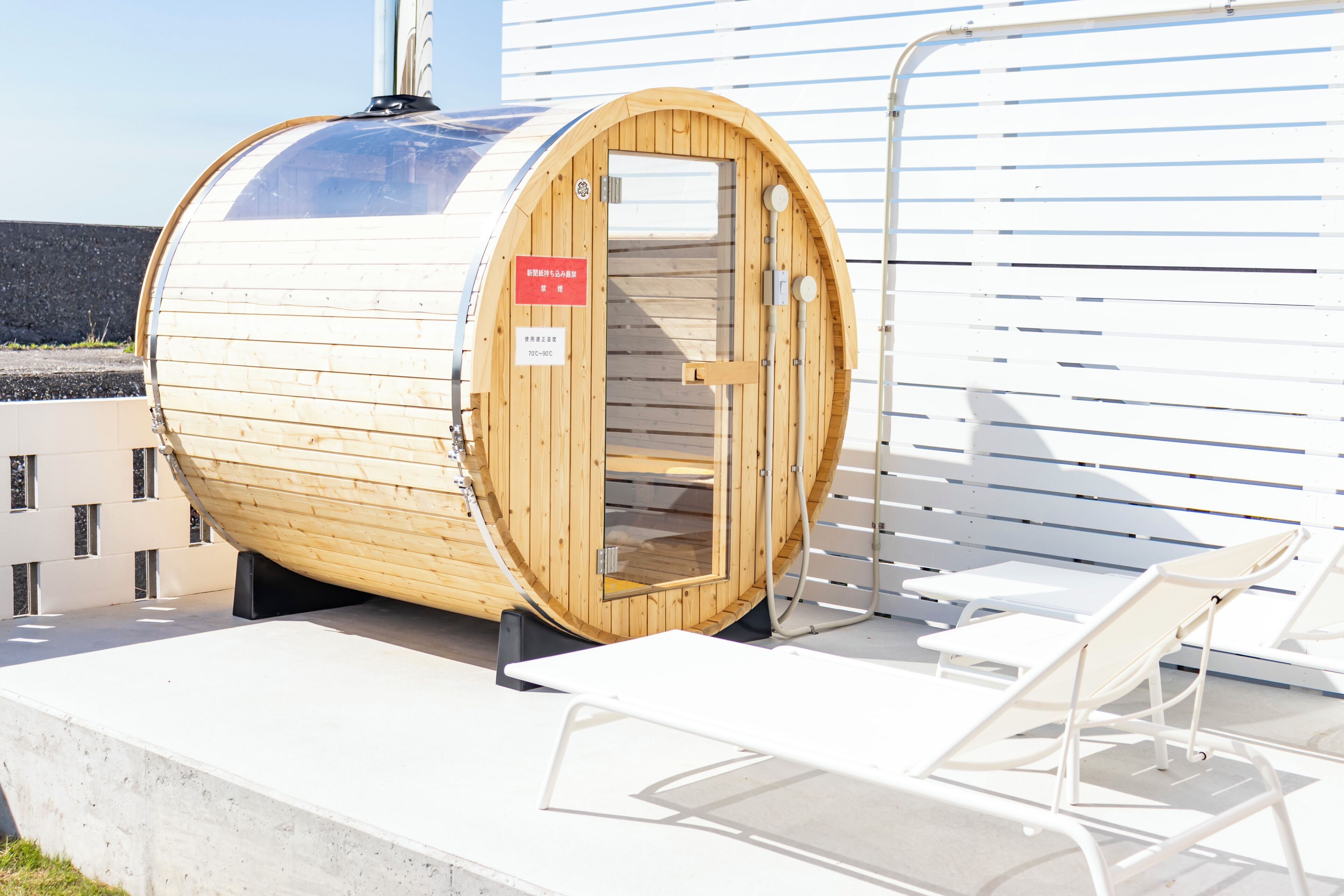 A sauna with a wooden exterior and a glass door, situated on a concrete platform with white lounge chairs nearby.