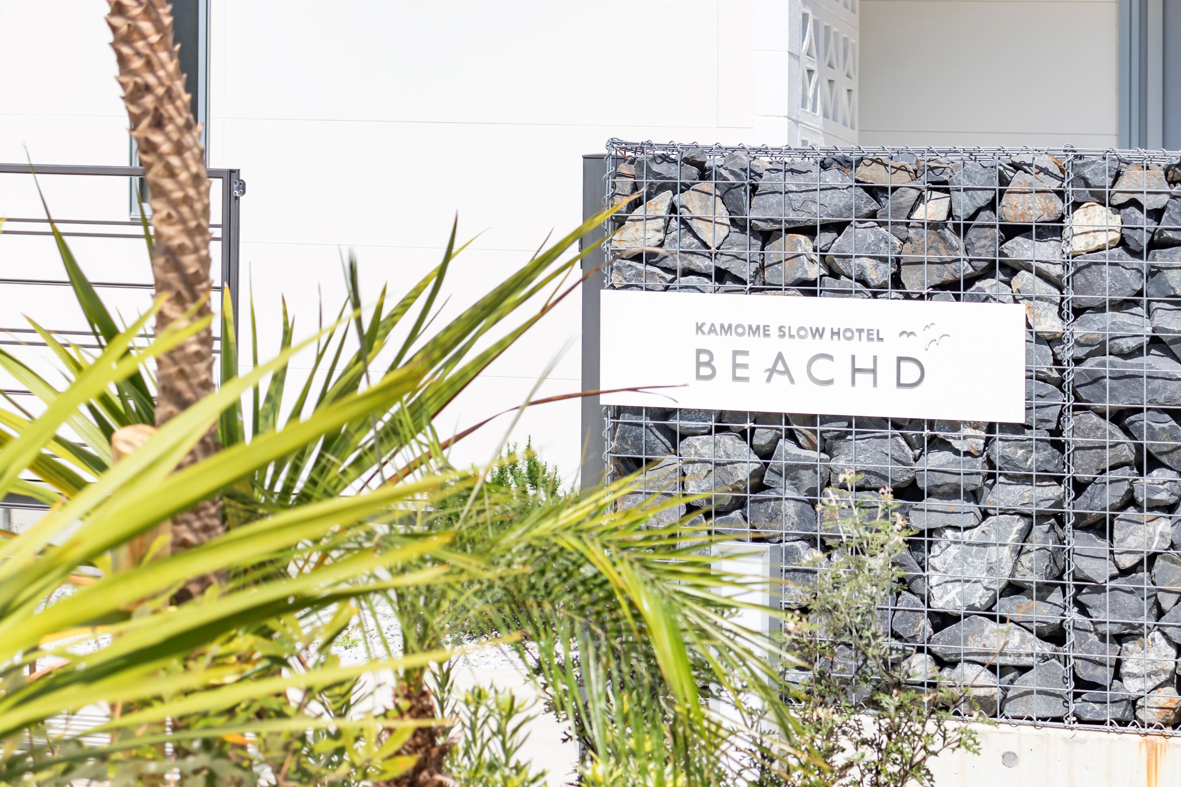 Une enseigne pour le Kamome Slow Hotel Beachd, avec un mur de pierres en arrière-plan et une végétation tropicale au premier plan.