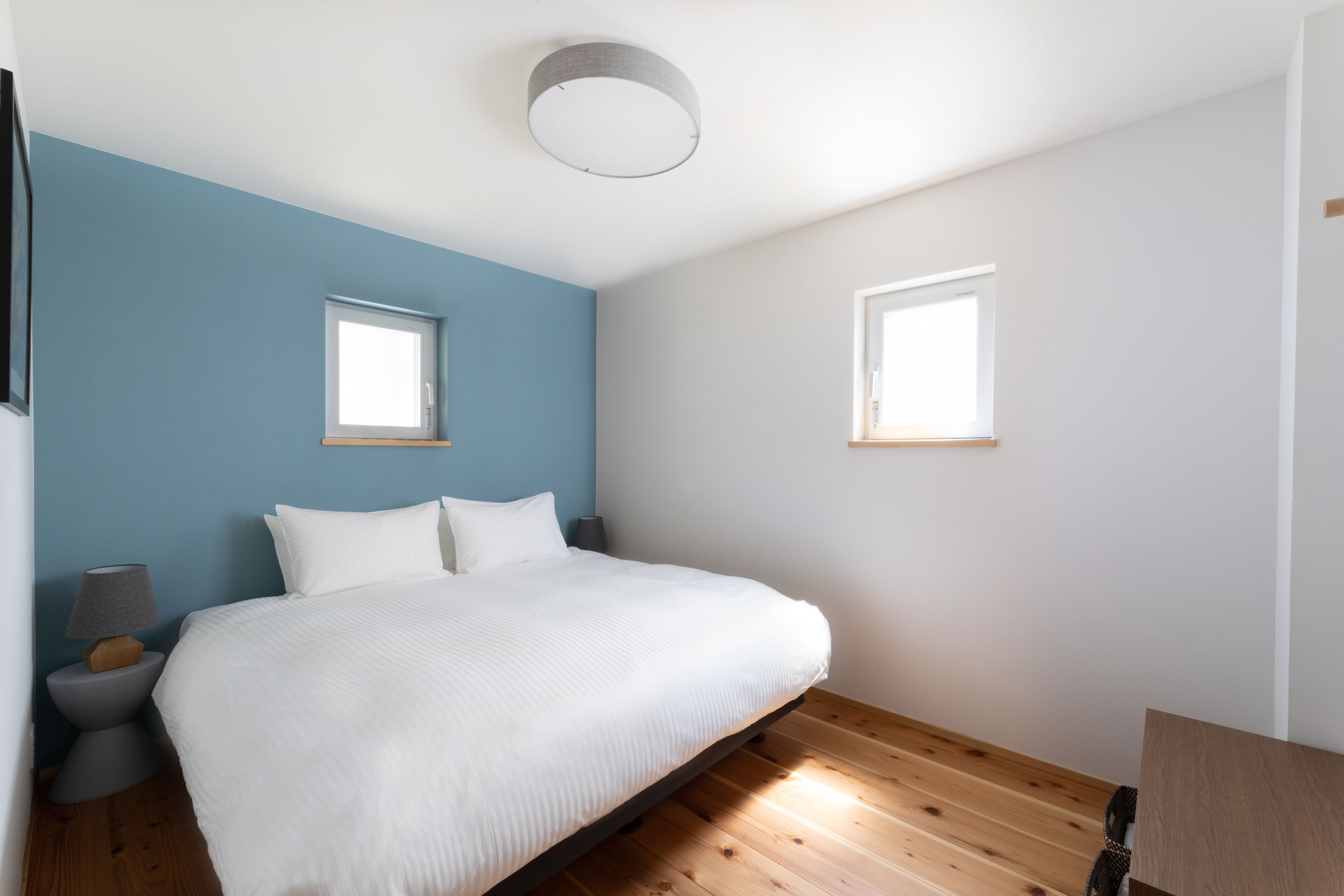 A bedroom with a bed, two windows, a ceiling light, and a wooden floor.