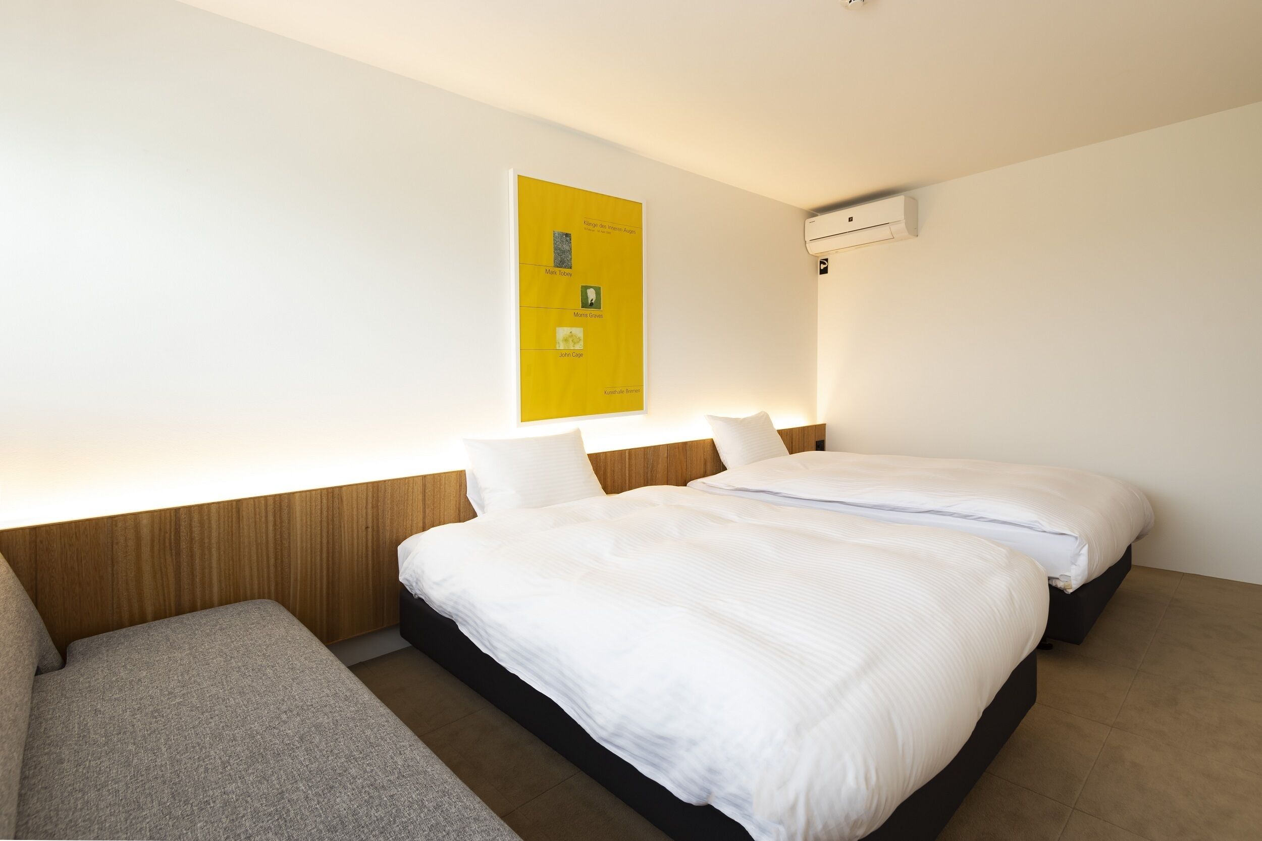 Une chambre d’hôtel avec deux lits, une tête de lit en bois, un mur blanc et une affiche jaune.