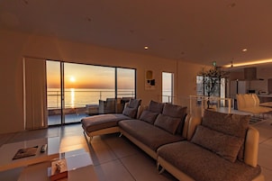 A modern living room with a large sectional sofa, a dining area, and a view of the sunset over the water.
