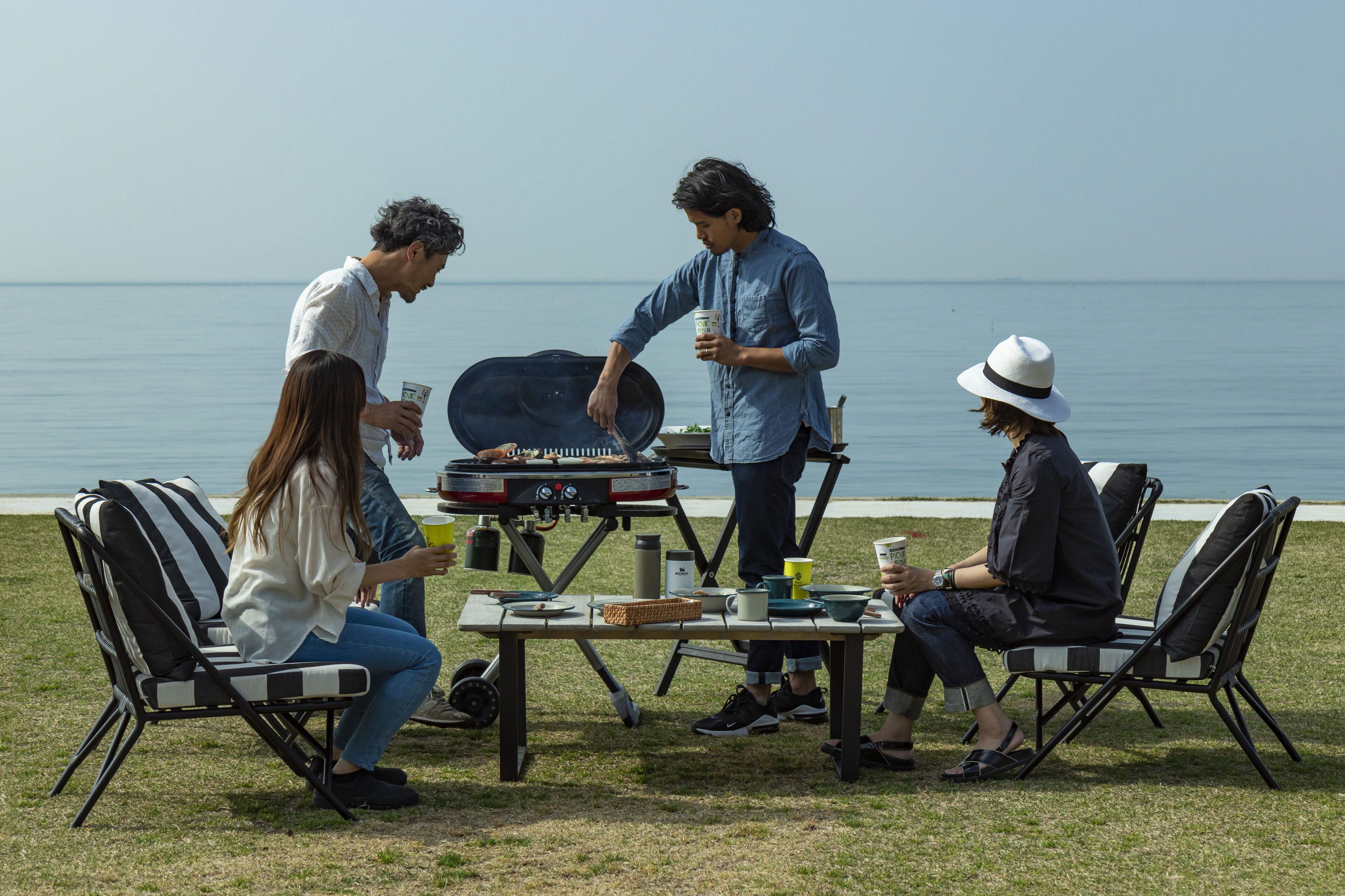 A group of people gathered around a portable grill outdoors with a view of the sea in the background.