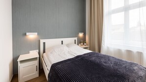 A neatly made bed with a white headboard, two bedside tables with lamps, a grey wall, and a window with curtains. - Victus Apartamenty - Malmo (Sopot)