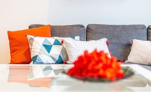 A sofa with multiple cushions, a glass table, and a red flower arrangement.