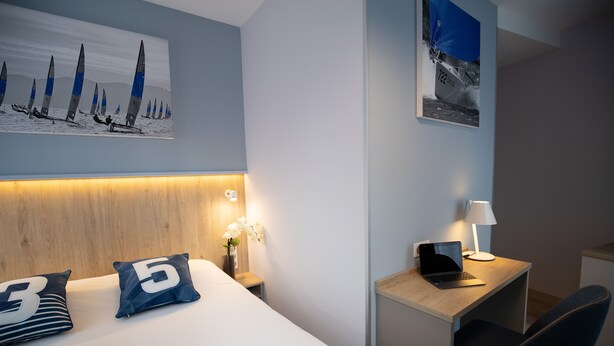 A hotel room with a bed, a desk, a chair, a nightstand with a lamp, and artwork on the walls. - Appartement des Marins (Saint-Malo)