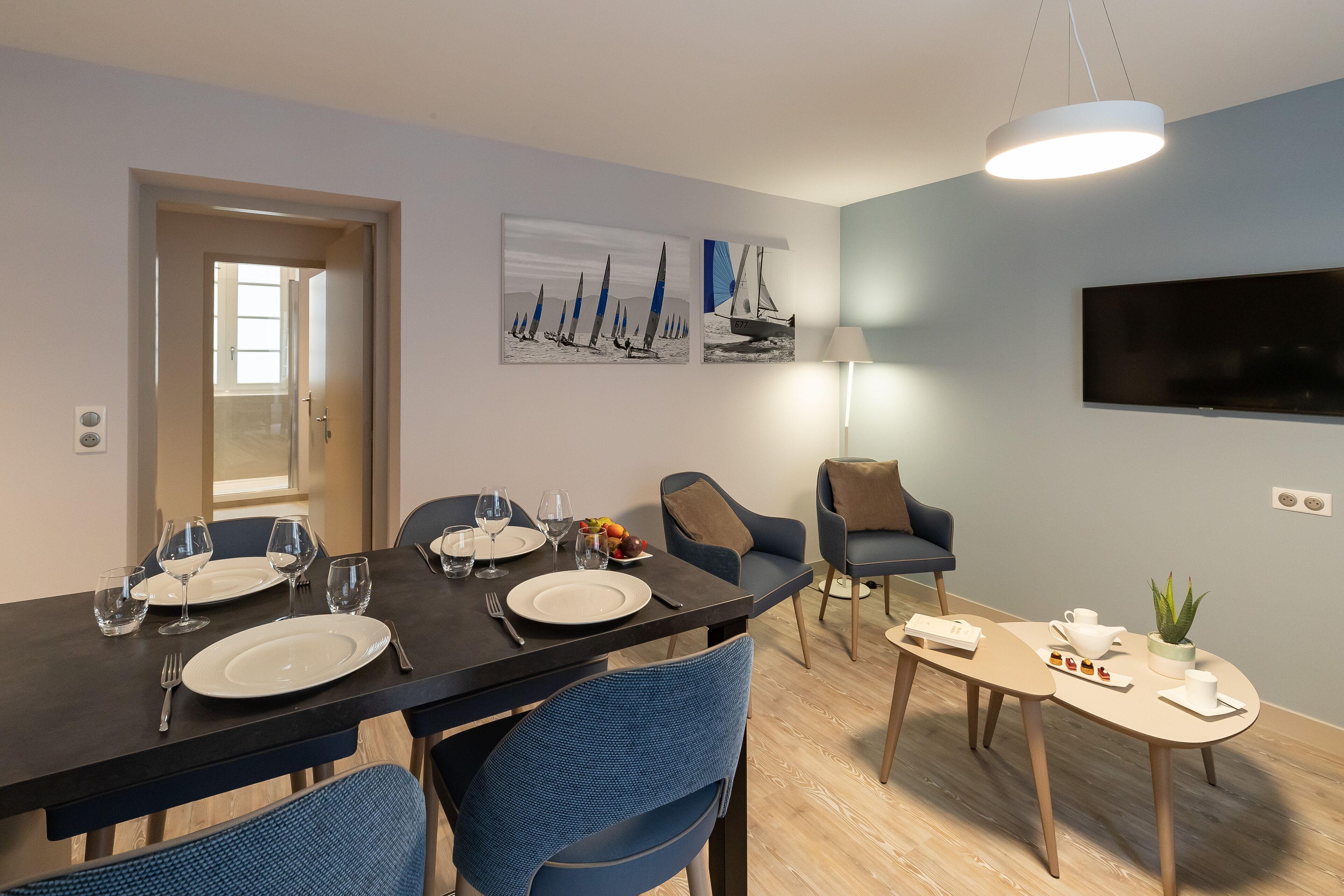 A dining area with a black table set for four, blue chairs, a small side table with a plant, and a TV mounted on the wall.
