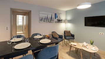 A dining area with a black table set for four, blue chairs, a small side table with a plant, and a TV mounted on the wall.