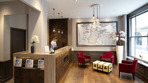 A reception area with a wooden reception desk, red chairs, a vintage suitcase, and a large framed artwork on the wall. - Appartement des Marins (Saint-Malo)