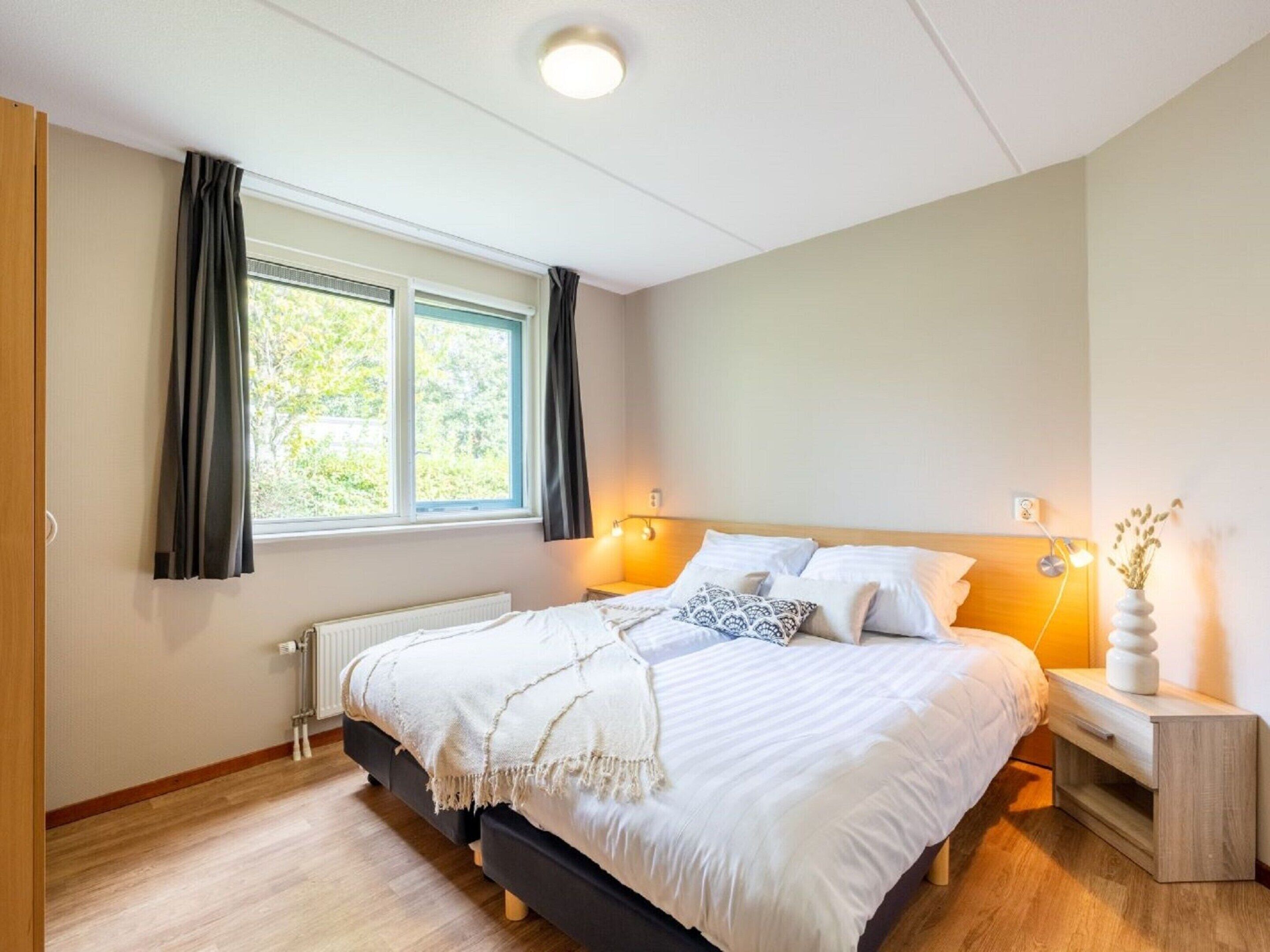 A bedroom with a bed, bedside tables, a window with curtains, a radiator, and a ceiling light.