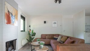 A modern living room with a fireplace, a brown sofa, two round tables, and decorative plants. - Woodland Bungalow w/ Pool (Epe)