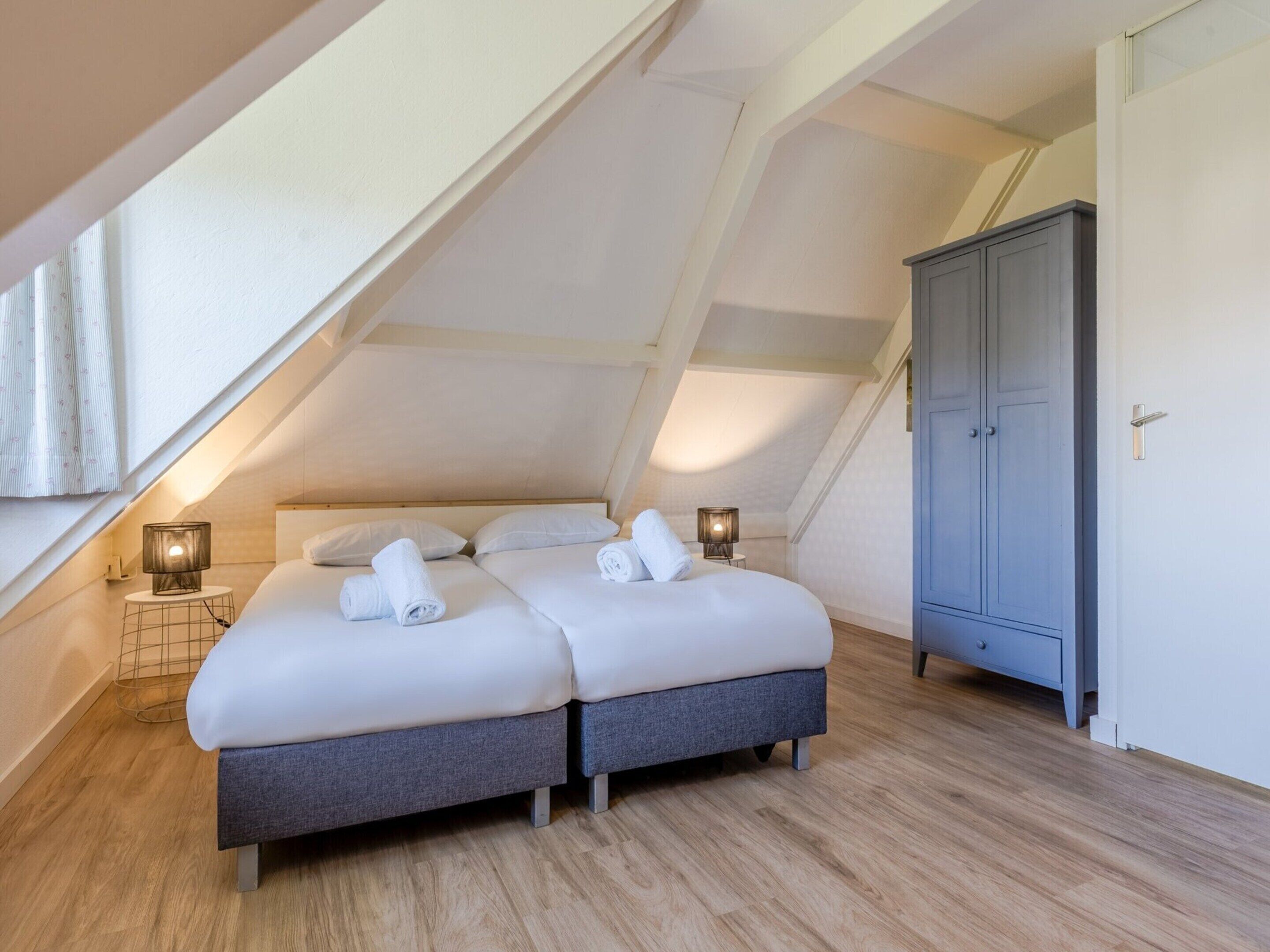 A two-bed room with a sloped ceiling, wooden flooring, a wardrobe, and bedside lamps.
