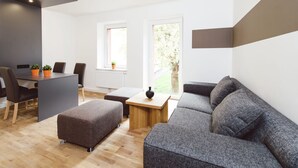 Un espacio de estar moderno con un sofá gris, una mesa de centro de madera y una zona de comedor con una mesa y sillas. Hay una ventana que proporciona luz natural y vistas a la vegetación exterior.