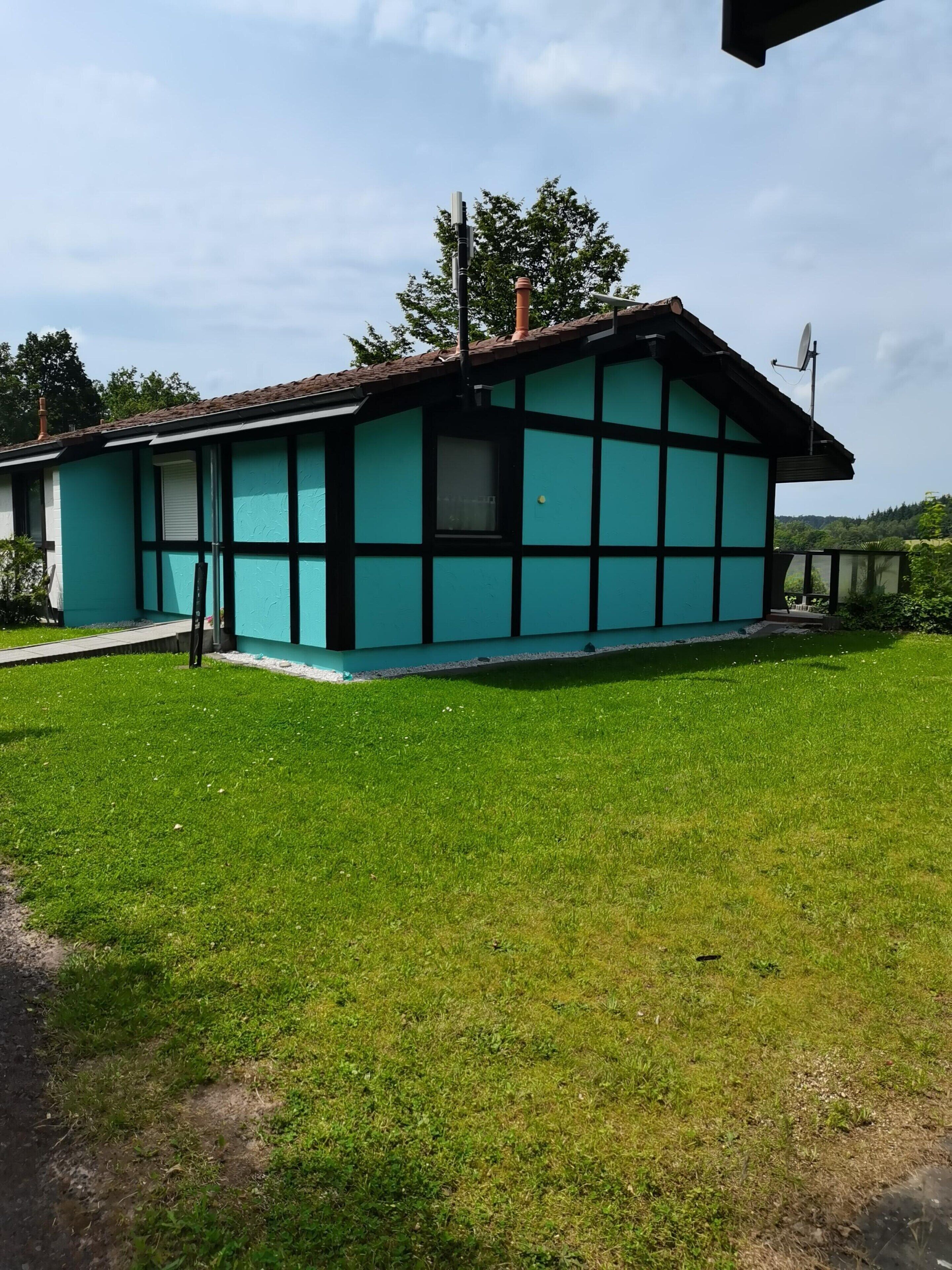 Ein eingeschossiges Gebäude mit türkis-schwarzer Fassade, einer sichtbaren Satellitenschüssel und einem Rasen.