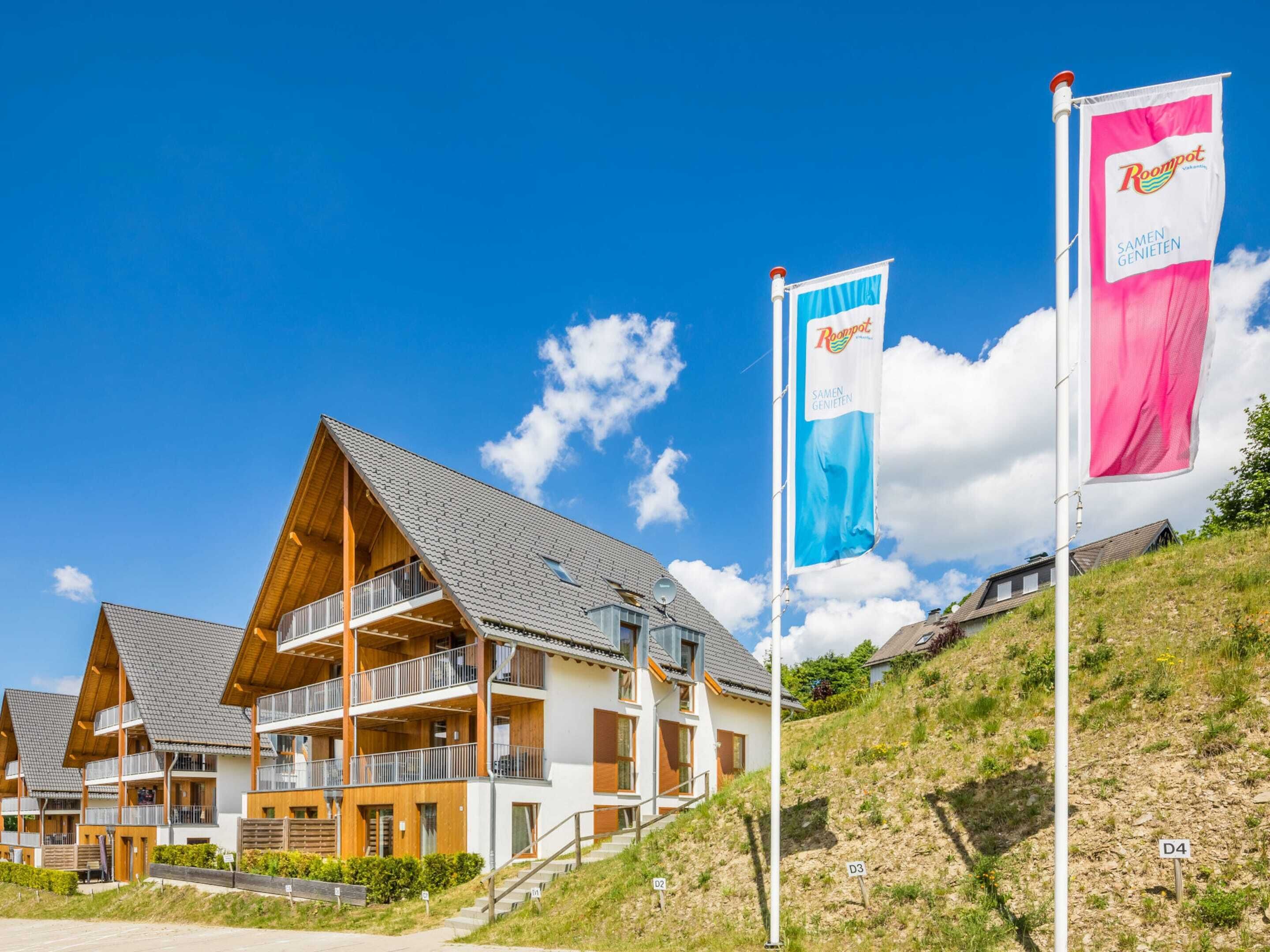 A modern building with multiple balconies and a sloped roof, surrounded by flags and a clear blue sky.