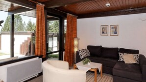 Ein gemütliches Wohnzimmer mit einem dunklen Sofa, einem weißen Sessel und einem kleinen Tisch. Zwei Fenster mit Vorhängen, eine Heizung und ein Deckenventilator sind vorhanden.
