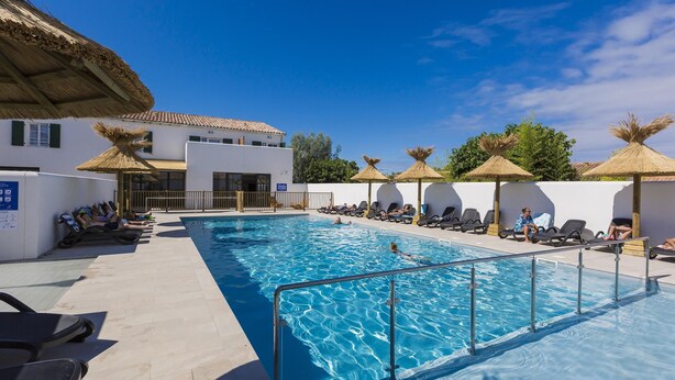 A swimming pool with lounge chairs and umbrellas, a building with a tiled roof, and a clear blue sky. - Nice Apartment on the Island of Ile de Re (Rivedoux-Plage)
